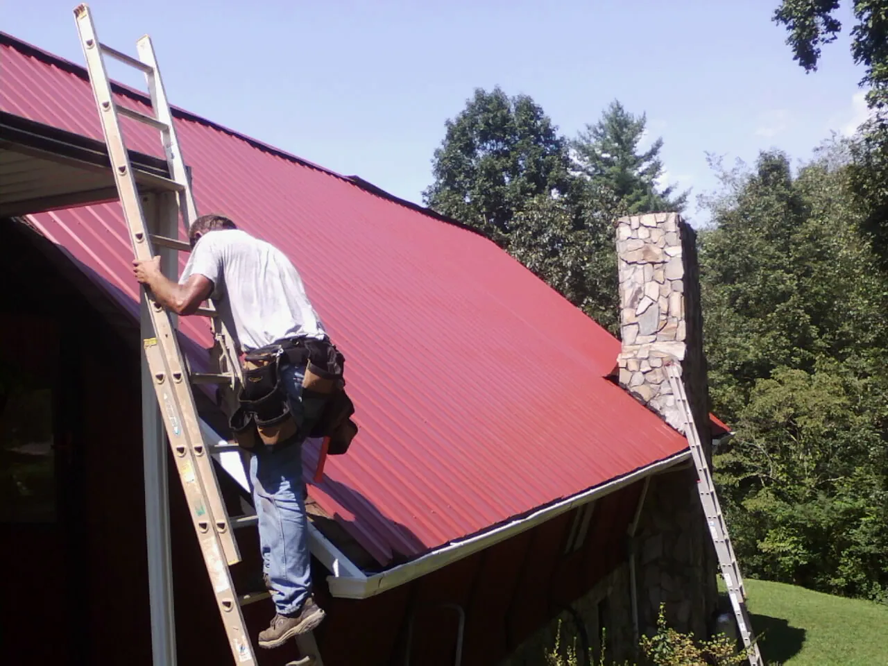 Roofer on a ladder beside a red metal roof and stone chimney — Soffit & Fascia Repair by Roofing Brothers in Deer Trail